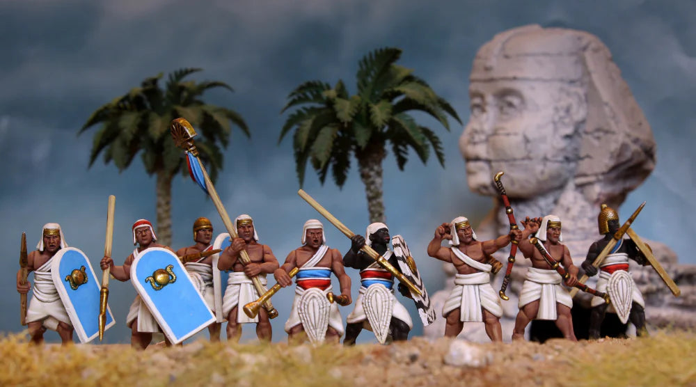 A set of miniature painted ancient Egyptian warrior figurines standing in a desert-like diorama with palm trees, a large stone head sculpture in the background, and a blue shield among their accessories.